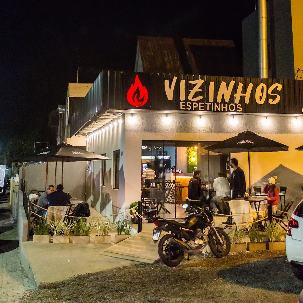 Área externa do Vizinhos Espetinhos ao entardecer, com iluminação aconchegante e mesas de madeira